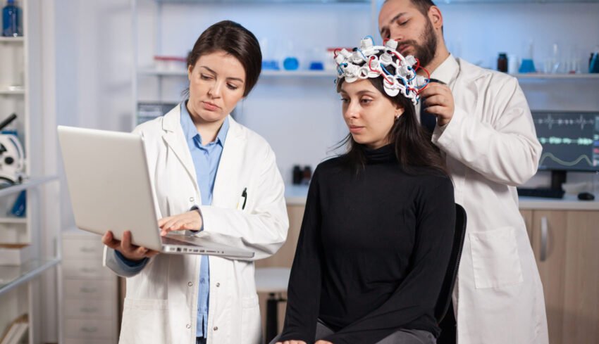 Specialist neurologist doctor taking notes on laptop asking patient's symptoms adjusting high tech eeg headset. Doctor researcher controlling EEG headset analyzing brain functions and health status.