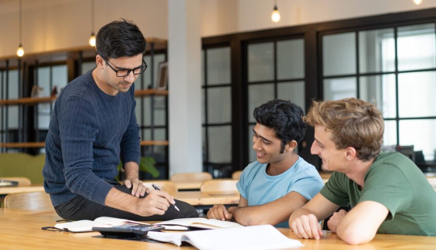 Serious teacher checking assignment of two students. Serious man in glasses pointing pen at notes of two cheerful guys. Education and internship concept